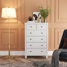 a white chest of drawers sitting next to a black chair and a lamp on top of a hard wood floor