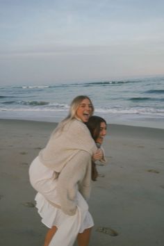 two women are walking on the beach with their arms around each other as they laugh