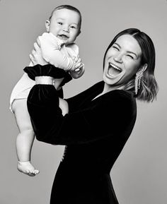 a woman holding a baby up to her face and smiling at the camera while wearing a black dress