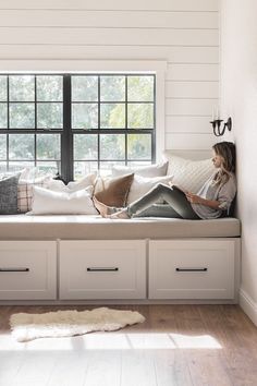 a woman sitting on top of a window sill next to a white bed with lots of pillows