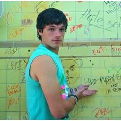 a young man standing in front of a wall with writing on it and pointing at something