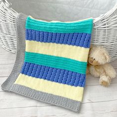 a teddy bear is sitting next to a knitted pillow on a white basket with a blue and yellow striped blanket