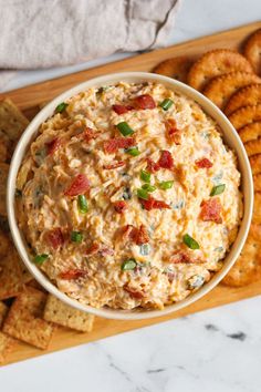 a bowl of dip surrounded by crackers on a cutting board with cheese and bacon