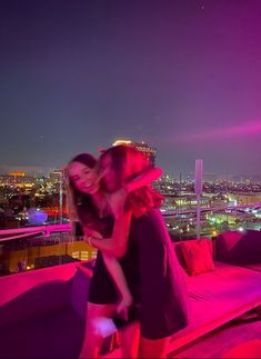 two women hugging each other on top of a building with city lights in the background