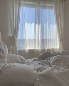 an unmade bed in front of a window with white drapes and curtains on the windowsill