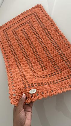 an orange crocheted blanket being held up by a person's hand on a white surface