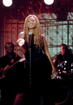 a woman standing in front of a microphone on stage with other people behind her and singing