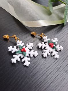 snowflake earrings with holly berries and bells are on a table next to a white ribbon