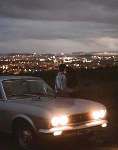 two people standing next to an old car in the dark with city lights behind them