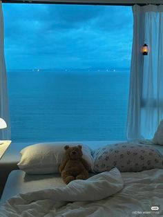 a teddy bear sitting on top of a bed in front of a window overlooking the ocean