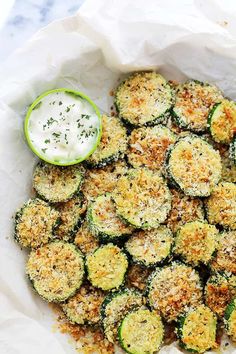 a white bowl filled with zucchini and cheese next to a small cup of ranch dressing