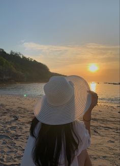 a woman sitting on top of a beach next to the ocean wearing a white hat