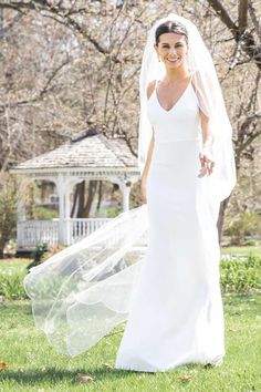 a woman in a white wedding dress and veil walking through the grass with trees behind her