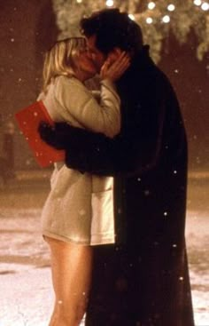 a man and woman kissing in the snow with lights behind them on a snowy night