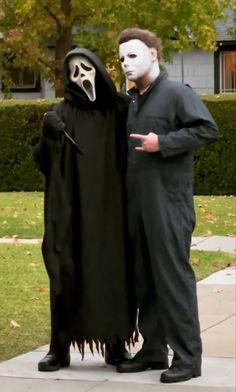 two people dressed up in halloween costumes standing next to each other on a sidewalk with trees and bushes behind them