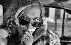 black and white photograph of a woman in sunglasses with her hand on her chin, leaning out the window of a car