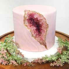 a pink and gold cake on a wooden table with greenery around the edges,