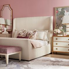 a white bed sitting next to a pink wall and a mirror on top of a dresser