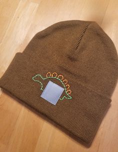 a brown beanie with a blue square in the center and a green dinosaur on it