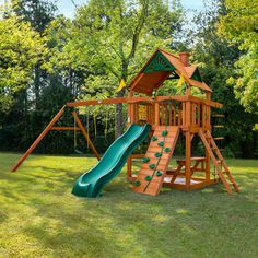 a wooden playset with a green slide and climbing frame in the grass next to trees