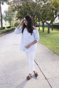 a woman in white is walking down the sidewalk with her hand on her head and looking at the camera