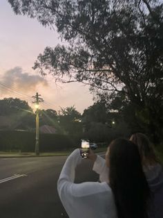 two women taking pictures with their cell phones on the side of the road at sunset