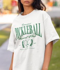 A fun graphic tee for the pickleballers! It's perfect for either the "social club" aesthetic or to wear when playing with your friends and teammates! For the "oversized" tee look, I recommend sizing up either 1 or 2 sizes from your typical day to day size! .: Made with 100% ring-spun cotton, a lightweight fabric (4.5 oz/yd² (153 g/m this unisex t-shirt feels like a bliss to wear all year round.  .: The classic fit with the crew neckline deliver a clean, versatile style that can match any occasion, whether it's formal or semi-formal.  .: All shirts feature a pearlized, tear-away label for total wearing comfort.  .: Made using ethically grown and harvested US cotton. Gildan is also a proud member of the US Cotton Trust Protocol ensuring ethical and sustainable means of production. This blank Tennis Tops With Graphic Print In Relaxed Fit, Sporty T-shirt For Pickleball In Summer, Sporty T-shirt For Summer Pickleball, Collegiate Crew Neck Tennis Tops, Casual Tennis T-shirt Relaxed Fit, Casual Graphic Print Top For Tennis, Casual Tennis T-shirt With Team Name, Crew Neck Tennis T-shirt With Team Name, Sporty White T-shirt For Pickleball