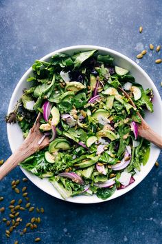 a white bowl filled with green salad and wooden spoons on top of blue surface