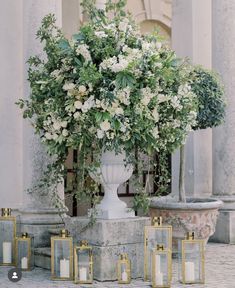 white flowers and greenery are in a vase on the ground next to some candles