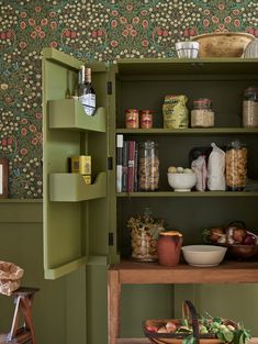 an open cabinet with many items on it in a room that has green walls and floral wallpaper