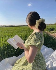 a woman in a green dress is reading a book on a white blanket near a field