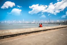 two people sitting on the edge of a wall looking out at the ocean and city