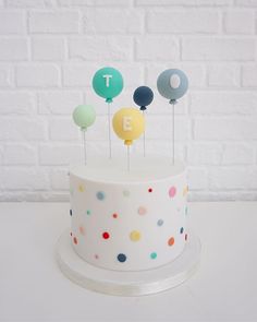 a white cake topped with lots of colorful dots and small balloons next to a brick wall