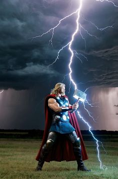 a man dressed as thor is standing in the grass under a lightning storm with his hammer