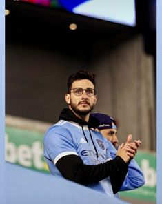 a man wearing glasses standing in front of a blue wall with his arms crossed and looking off to the side