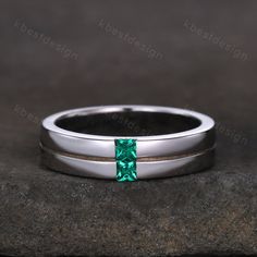 a white gold wedding band with a square cut emerald colored stone in the center, on top of a rock