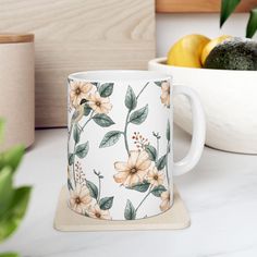 a white coffee mug sitting on top of a counter next to a bowl of fruit