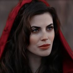 a woman with dark hair and blue eyes wearing a red cloak over her head is staring at the camera