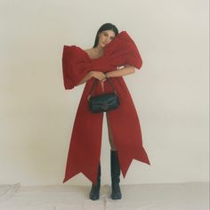 a woman wearing a red coat and holding a black handbag in front of a white wall