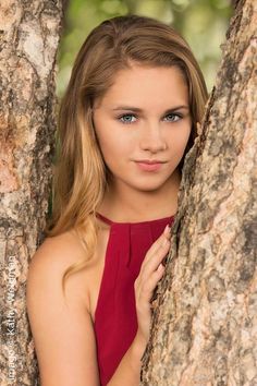 a beautiful young woman in a red dress leaning against a tree