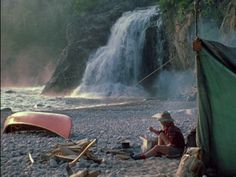 a man sitting on the ground next to a tent near a waterfall with a kayak