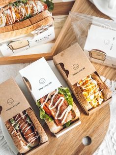 three sandwiches in boxes sitting on top of a wooden table next to cups and saucers