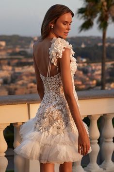 a woman in a short white dress standing on a balcony looking at the city behind her