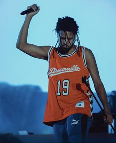 a man with dreadlocks and an orange shirt holding a microphone in his right hand