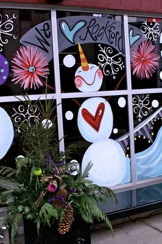 a window decorated with snowmen and flowers on the side of a building, in front of a potted plant