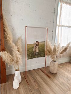 two vases with dry grass in them next to a photo on the wall and an easel