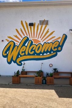 a welcome sign painted on the side of a building with potted plants in front