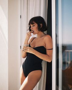 a woman in a black swimsuit leaning against a window with her hand on her lip