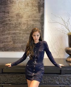 a beautiful young woman standing in front of a table with a vase on top of it