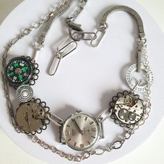 I have recycled three vintage mechanical watches for this Steampunk-inspired necklace adding a brass-with-white calendar watch dial, a reclaimed metal utility button ["guaranteed authentic"], bracelet sections from another antique wristwatch, a contemporary button compass as another contrast dial and reclaimed lengths of chain. To complete the total length of approximately 16.00 inches I added paperclip chain. It has been photographed at 14.00 inches. This necklace is listed in my shop twice as Vintage Round Compass Jewelry, Vintage Silver Watches With Compass Design, Vintage Adjustable Stainless Steel Watch, Vintage Stainless Steel Watch With Adjustable Fit, Vintage Stainless Steel Adjustable Watch, Vintage Metal Round Jewelry And Watches, Vintage Metal Pocket Watch Nickel Free, Retro Soldered Metal Jewelry, Vintage Stainless Steel Jewelry With Compass Design